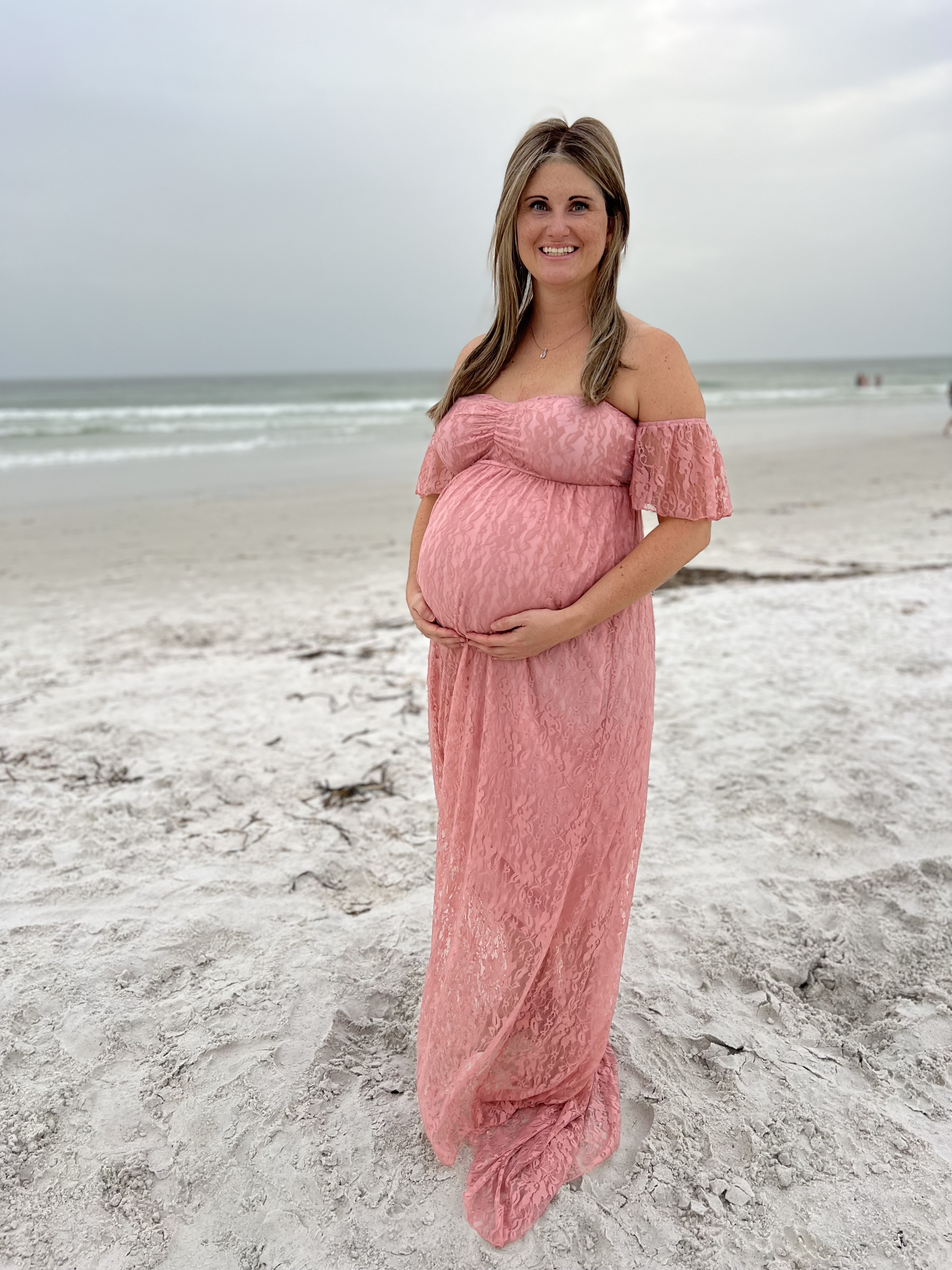 Holly young white woman with long hair and very pregnant wearing a pink off the should lace dress standing on a beach