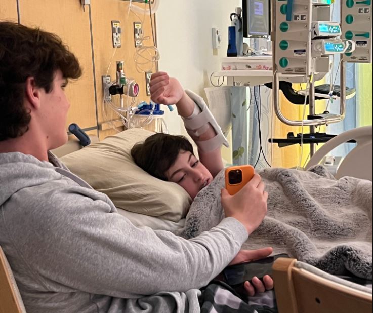 young white teen boy with dark hair lying in a hospital bed with his arm extended over his head looking at an orange phone being held by another teen boy