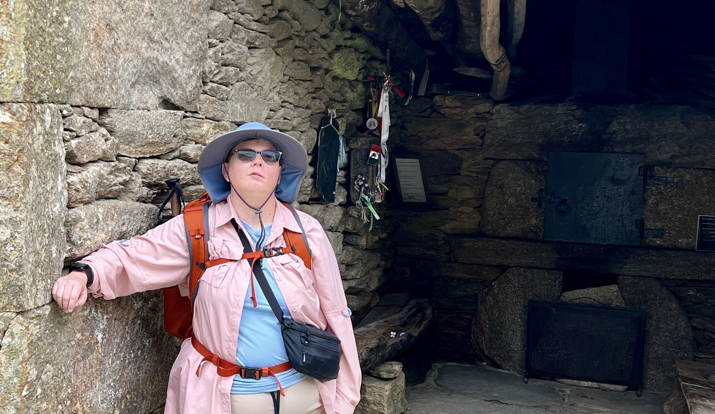 Kendra stands against a stone wall, wearing a pink shirt, hiking gear, and a hat.