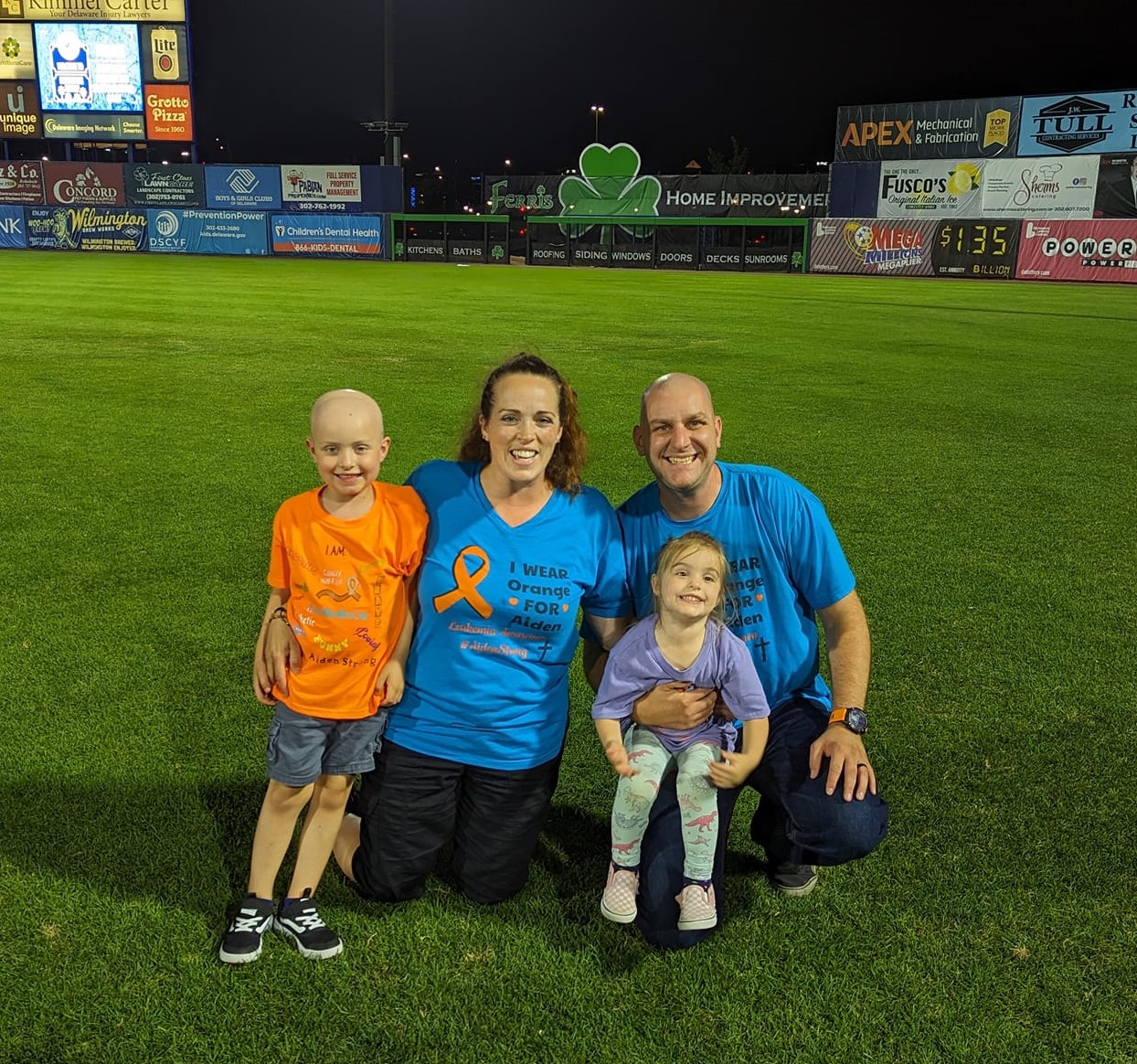 little white boy who is bald wearing an orange t-shirt and blue shorts next to his mom sister and dad wearing blue and orange tshirts on a ball field