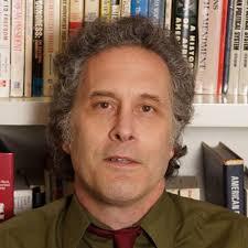 middle aged white man with graying sideburns and a green shirt and red tie sitting in front of a bookcase