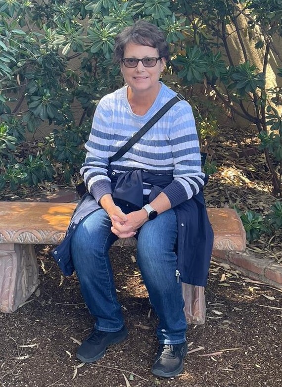 middle aged white woman short brown hair sitting on a bench and wearing a blue striped shirt and jeans with a jacket tied around her waist