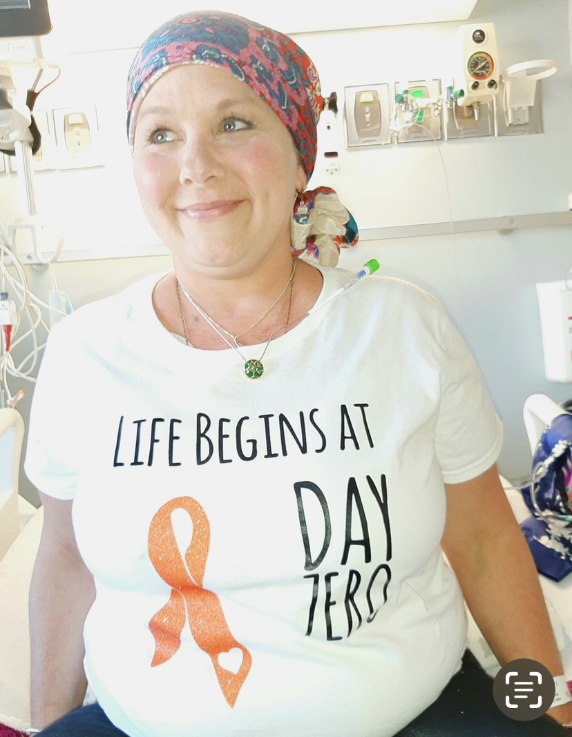 middle aged white woman wearing a colorful headscarf and white t-shirt with an orange ribbon on it sitting on a hospital bed