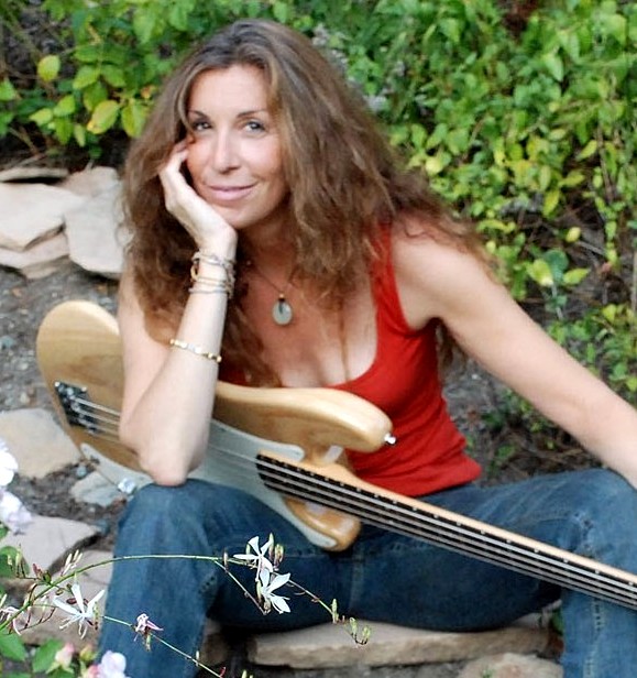 middle aged white woman with long brown hair wearing and orange tank and jeans with a guitar on her lap sitting in a garden