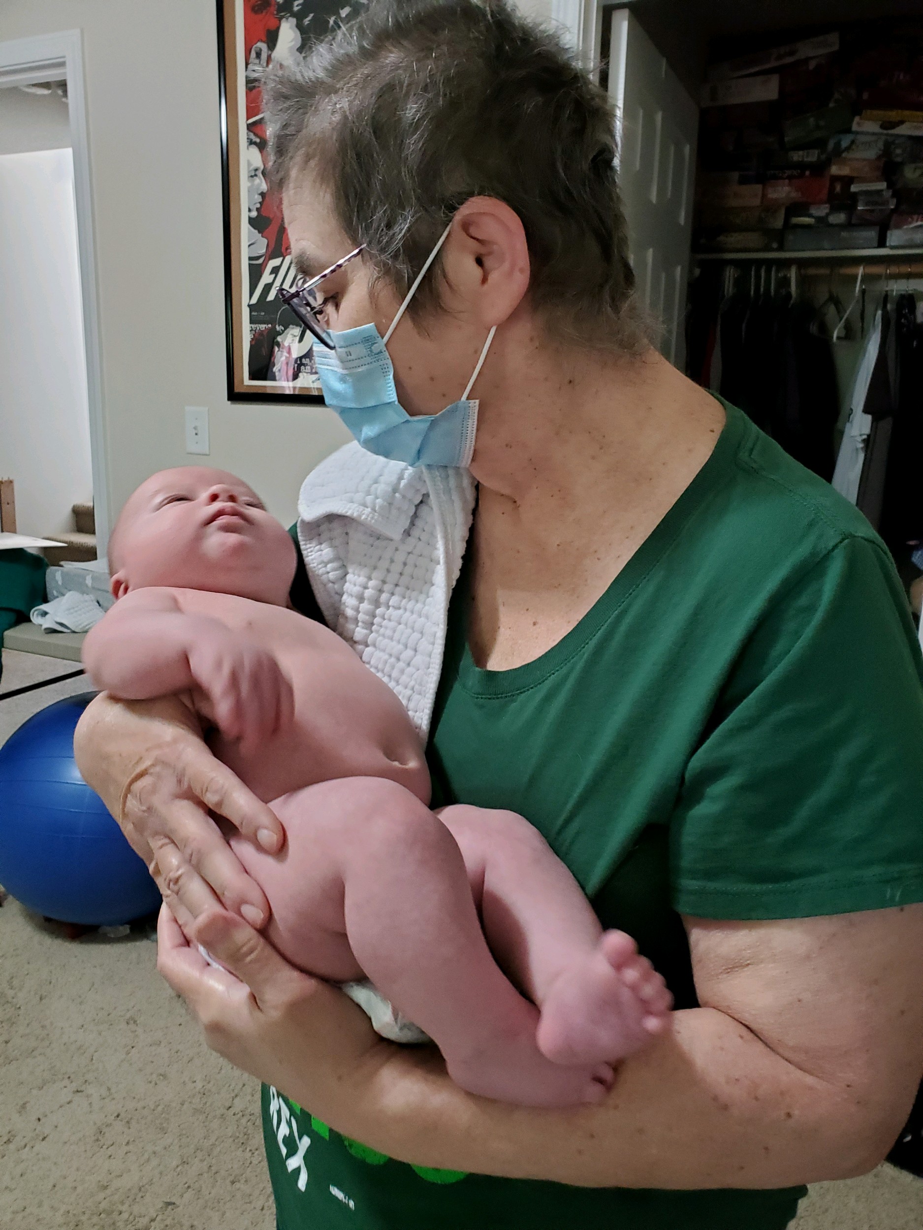 middle aged white woman with short hair and glasses wearing a face mask and a green shirt holding a baby