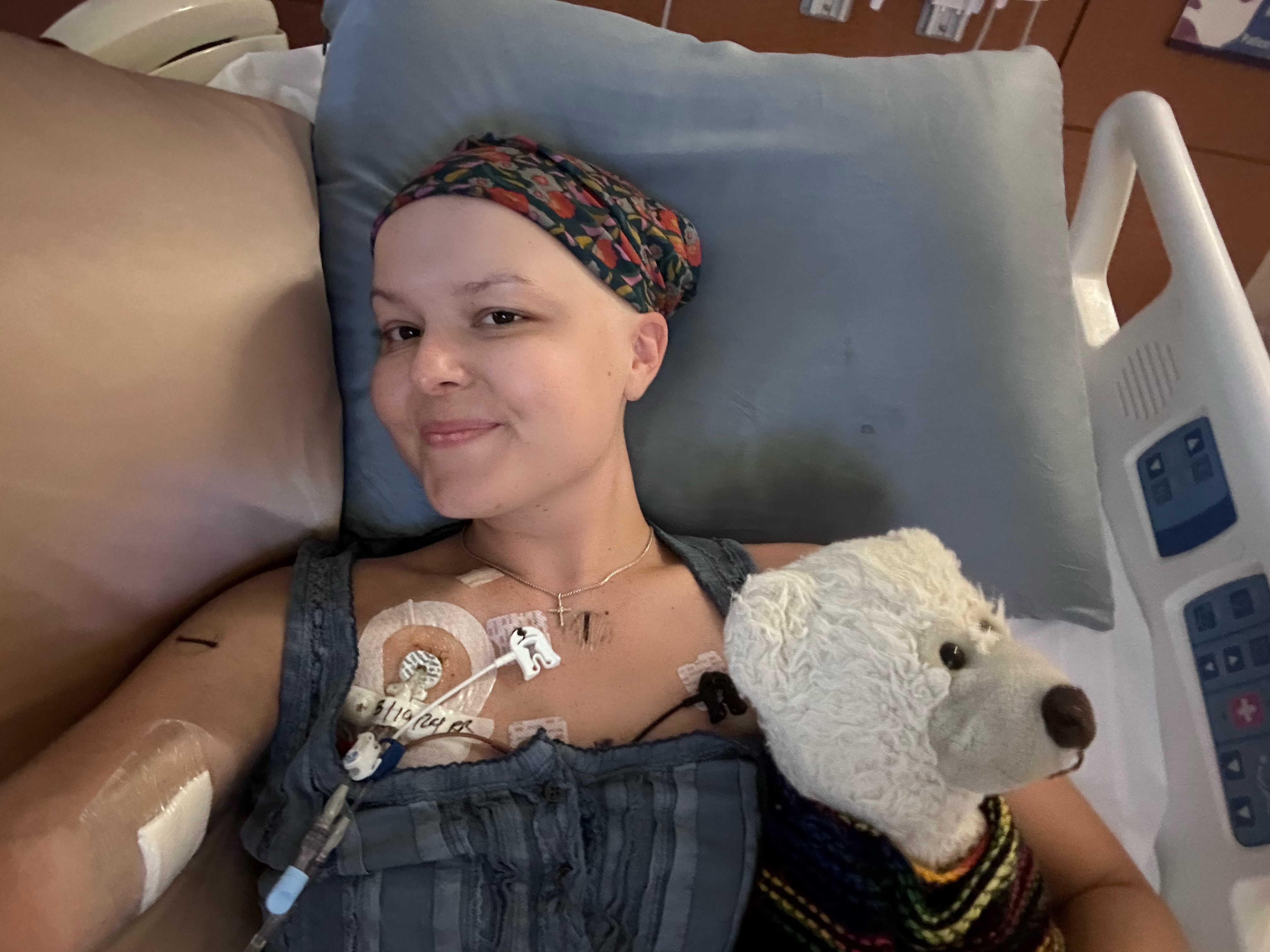 young white woman wearing a head scarf and gray top lying in a hospital bed with a stuff white bear