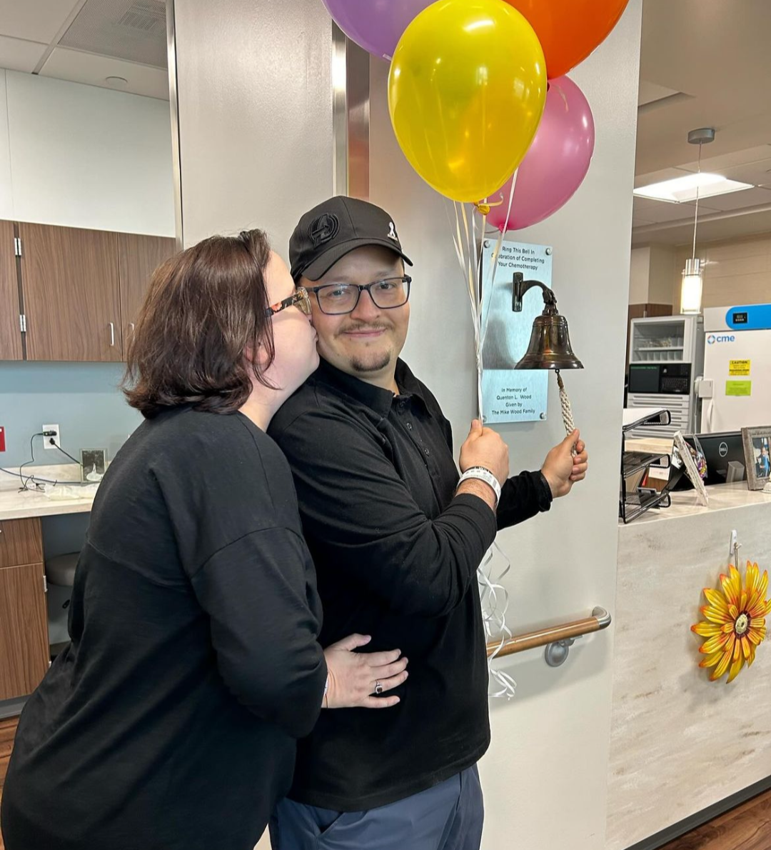 Hispanic couple both dressed in black se is kissing him as he rings the bell and holds balloons