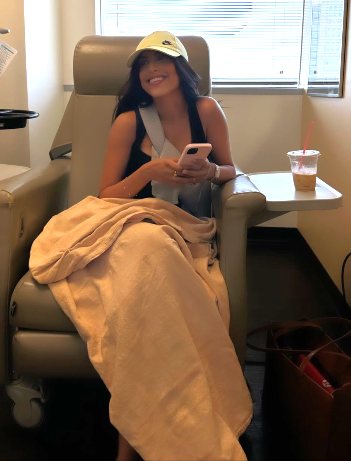 young ethnic girl sitting in a hospital chair wearing a yellow ball cap and a black sleevelss tshirt holding a pink phone with a blanket on her lap