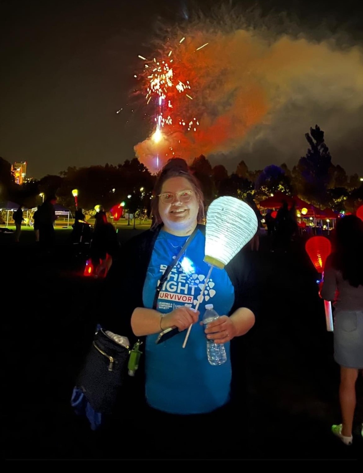 young white girl with her hair pulled up with glasses wearing a black jacket and blue tshirt carrying a white balloon at LTN with fireworks in the background