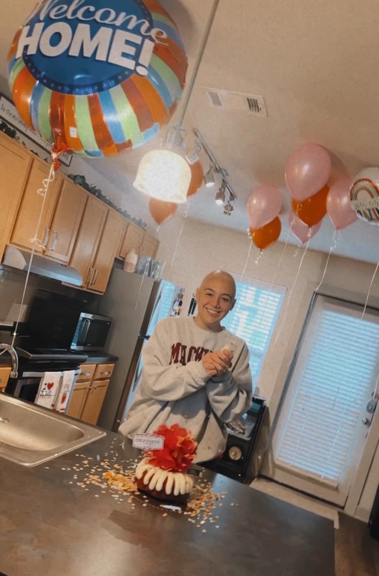 young white woman bald with a nose ring wearing a sweatshirt and standing in a kitchen with balloons and a bundt cake