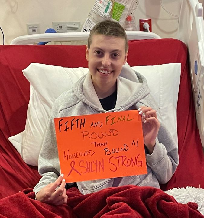 young white woman in a hospital bed with short hair wearing a gray hoodie and holding an orange sign about fifth round of chemo