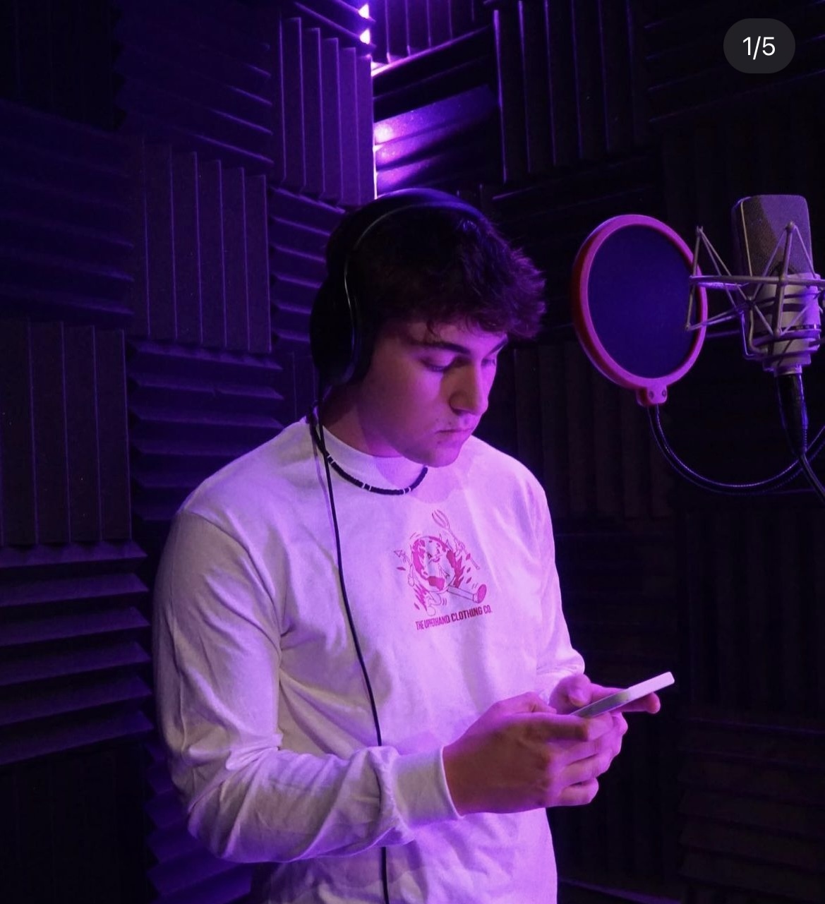 Marko young white male with curly dark hair in a recording studio wearing headphones and a puka shell neclklace and a white long sleeve tshirt with a pink logo also holding a phone