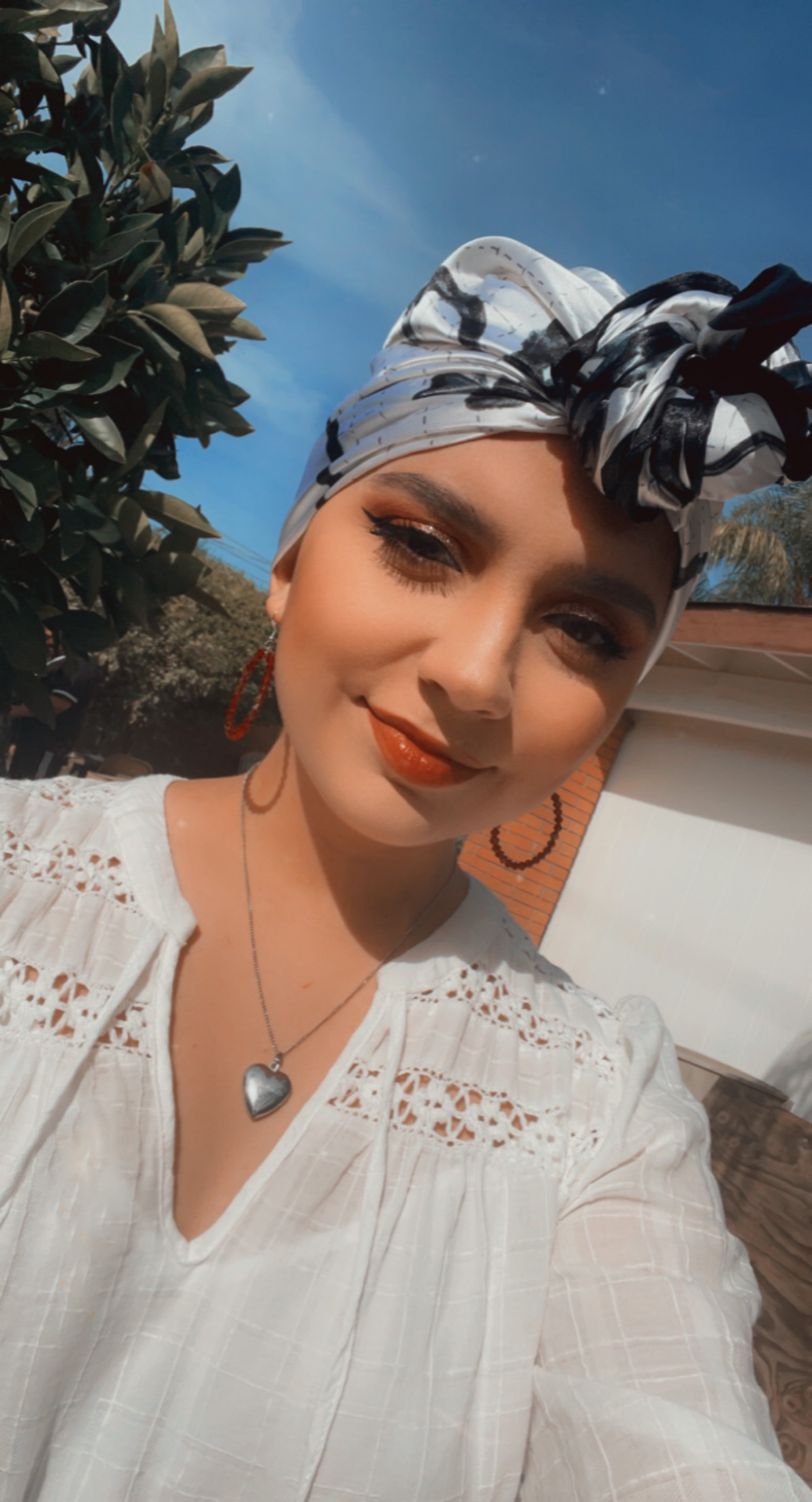 Yesenia young hispanic woman with blue and black headscarf on hoop earrings and a heart necklace wearing a white lacey blouse