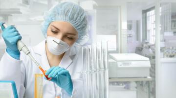 Researcher wearing a hair net, N95 mask and gloves injects a liquid into a vial. 