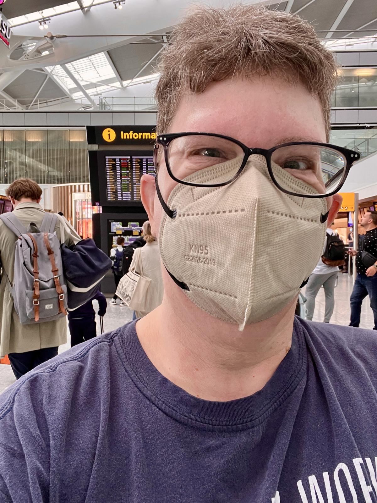 Kendra wears a respirator mask in an airport terminal. Behind her is a departures board.