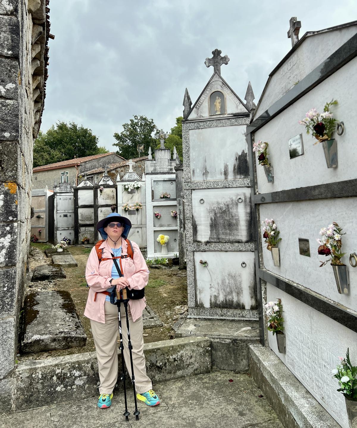 Kendra stands in a historic graveyard on the Santiago de Compostela pilgrimage route. She wears a hat and hiking backpack and is carrying trekking poles.